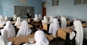 A classroom of young women sit at desks and focus on a female teacher in the front of the classroom. 