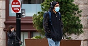 A male student walks with a backpack on, mask, and headphones